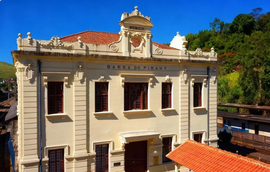 Vista frontal do Centro Cultural Rosemar Muniz Pimentel localizado na antiga estação ferroviária de Barra do Piraí. O edifício histórico possui cor bege com detalhes em branco e a inscrição Barra do Pirahy no topo. É possível ver parte dos trilhos ferroviários nas laterais sob um céu azul limpo.