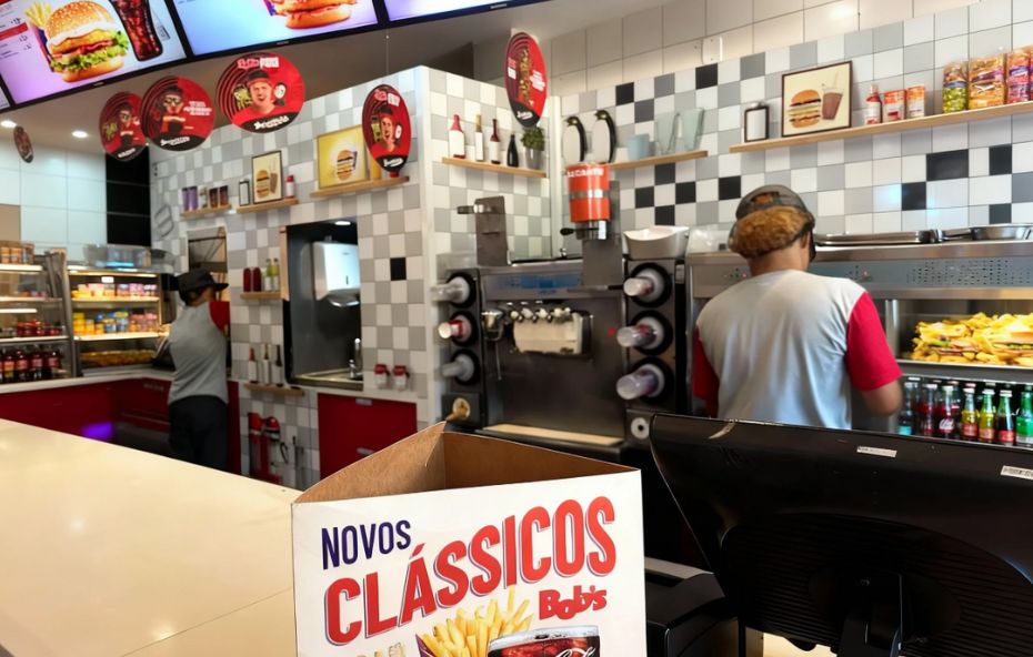 Visão interna de uma lanchonete com balcão branco em primeiro plano, funcionários uniformizados trabalhando ao fundo, paredes com azulejos quadriculados em branco e cinza, máquinas de sorvete e telas de cardápio digital suspensas.