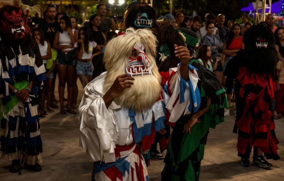 Fotografia noturna em plano médio mostrando três foliões caracterizados como palhaços de Folia de Reis, com máscaras artesanais expressivas e roupas coloridas de fitas, enquanto o público observa ao fundo na Praça da Estação.