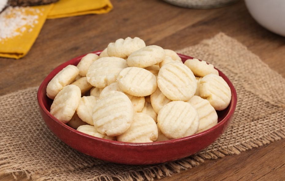 Tigela vermelha com biscoitos amanteigados claros e arredondados dispostos sobre um pano rústico em uma mesa de madeira.