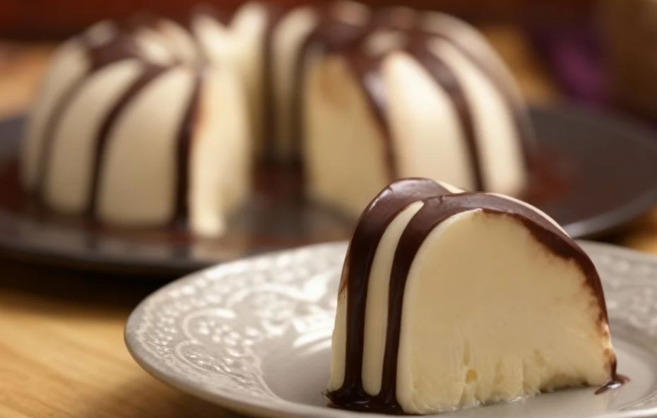 Fatia de bolo de sorvete branco com calda de chocolate escorrendo em destaque sobre prato de louça clara com o restante do bolo ao fundo.