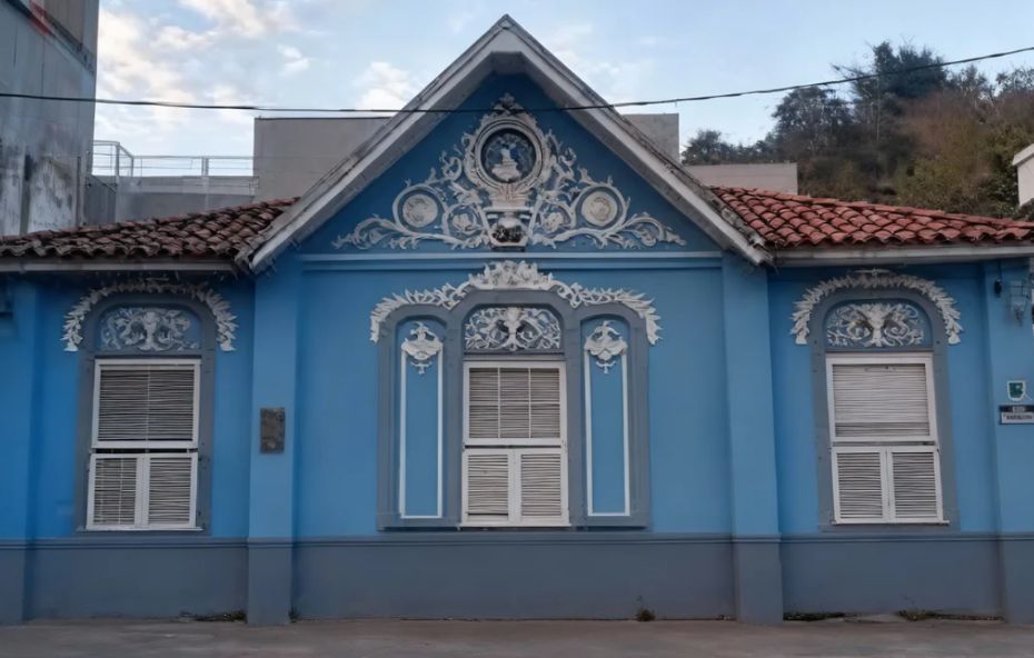 Fotografia da fachada de uma casa estilo chalé na cor azul claro com detalhes ornamentais em branco. O prédio possui um frontão triangular central elevado contendo um medalhão e guirlandas em relevo acima das janelas brancas com venezianas.