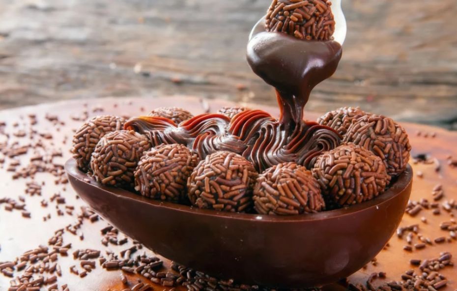 Uma metade de casca de ovo de chocolate recheada com diversos brigadeiros redondos cobertos com granulado escuro. Uma colher de metal despeja uma calda de chocolate espessa e brilhante sobre os doces em um cenário rústico de madeira.